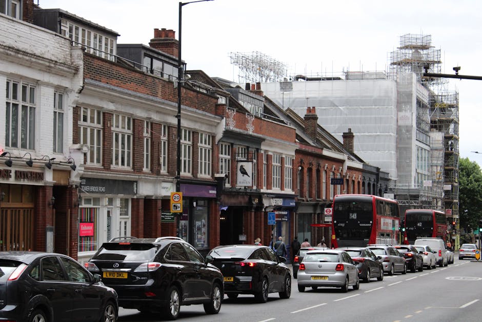 A busy street scene showing a row of brick and painted shops and businesses with large windows, some with awnings, located along a pavement. Several cars, including black, grey, and white vehicles, ar